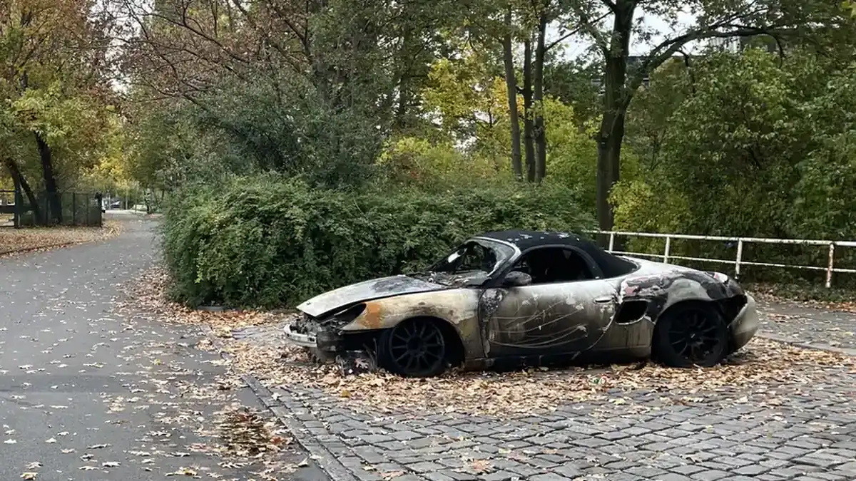 Burned-out silver convertible sports car parked on cobblestones beside a hedge, its paint scorched and windshield gone