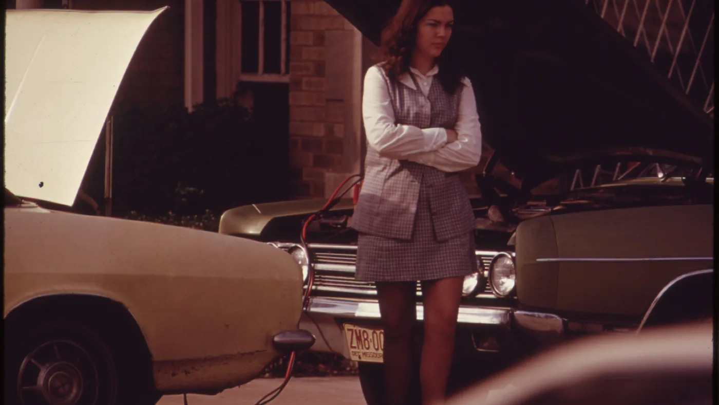 A woman waits beside two cars with their hoods open and jumper cables connected between them, outside a brick building