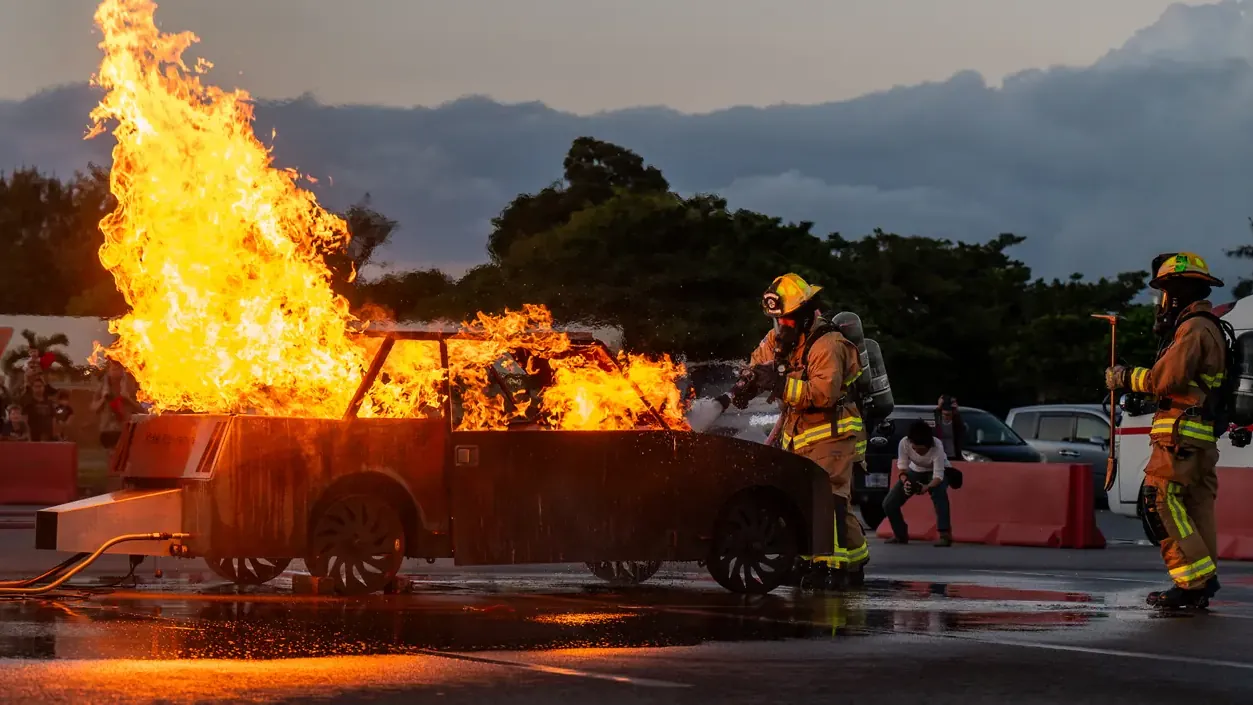 Firefighters in turnout gear spraying water on a burning car-shaped training prop at dusk during a fire-safety demonstration