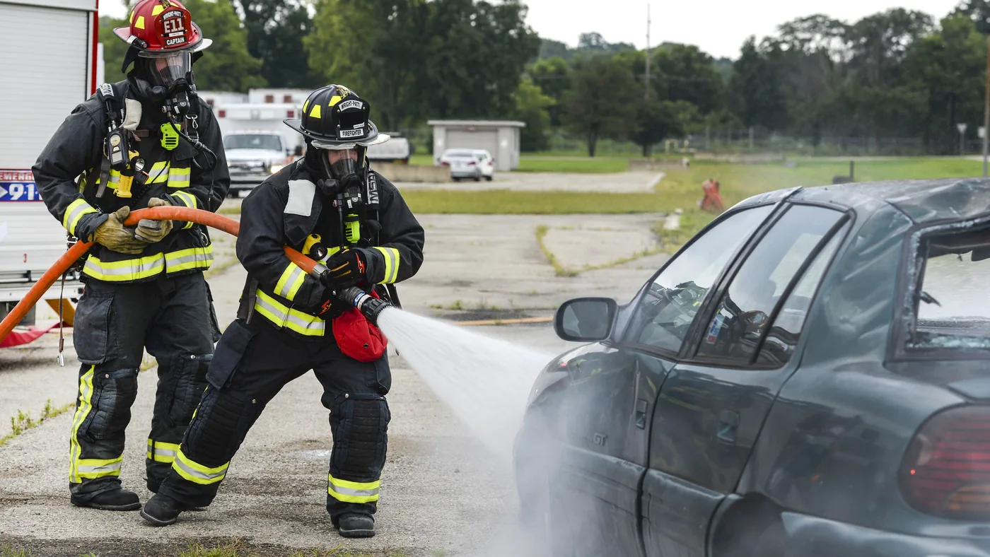 Two firefighters in breathing gear aiming a hose at a dark sedan during a vehicle fire training exercise