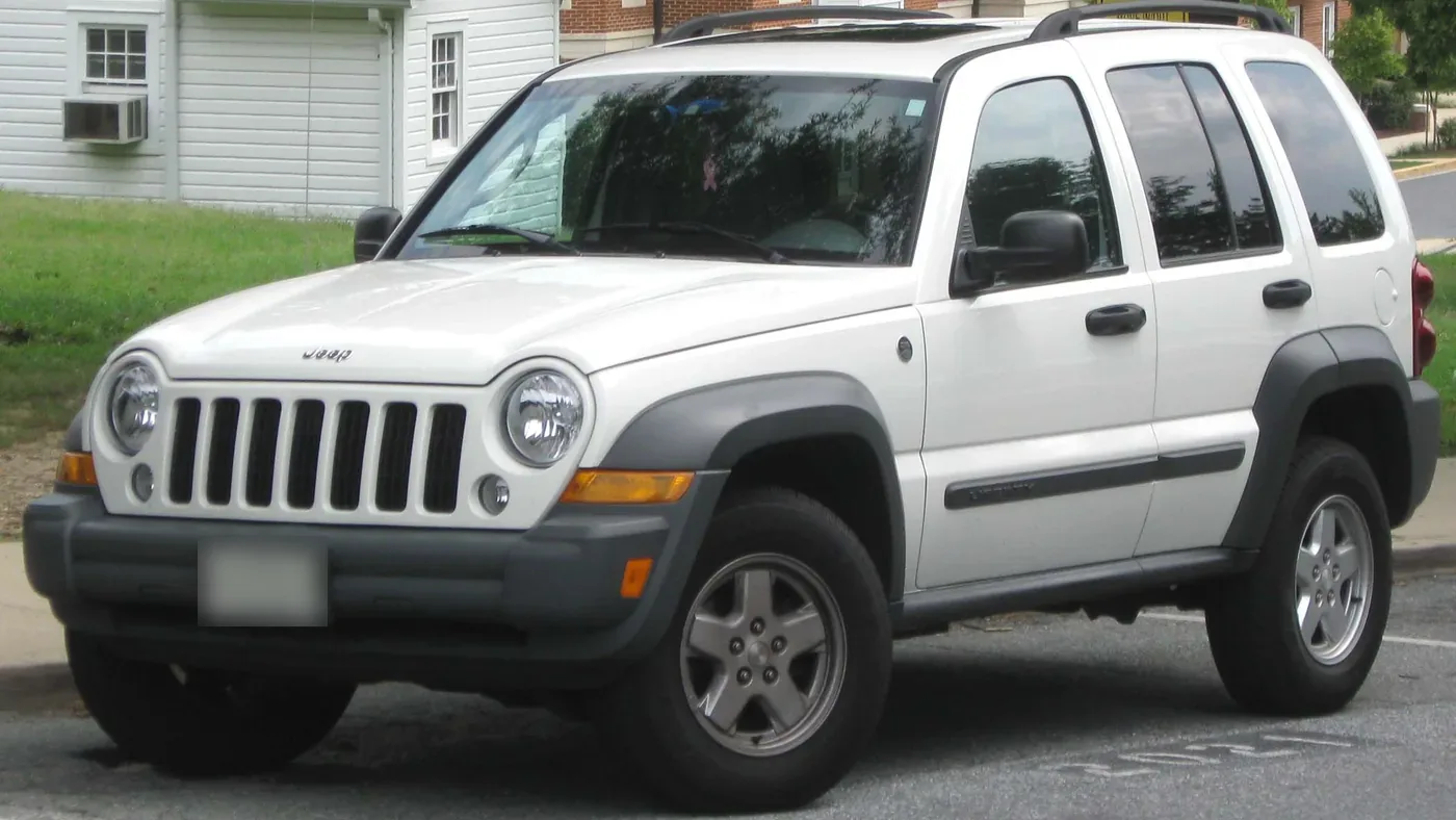 White 2005–2007 Jeep Liberty SUV parked at a curb beside a lawn and a white house, seen from the front left
