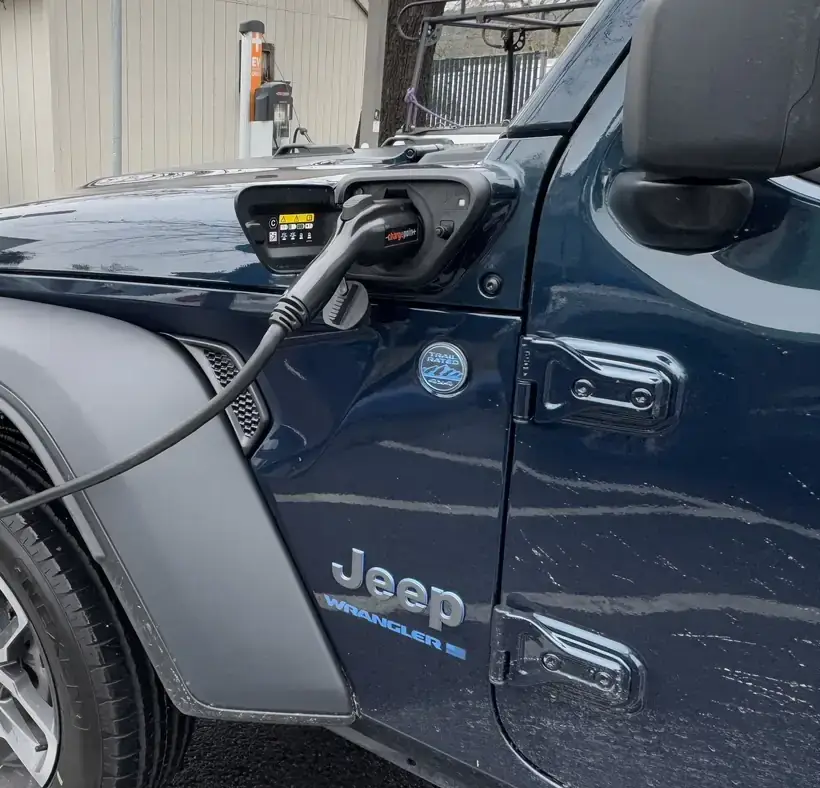 Blue Jeep Wrangler 4xe plug-in hybrid with a charging cable plugged into the port on its front fender