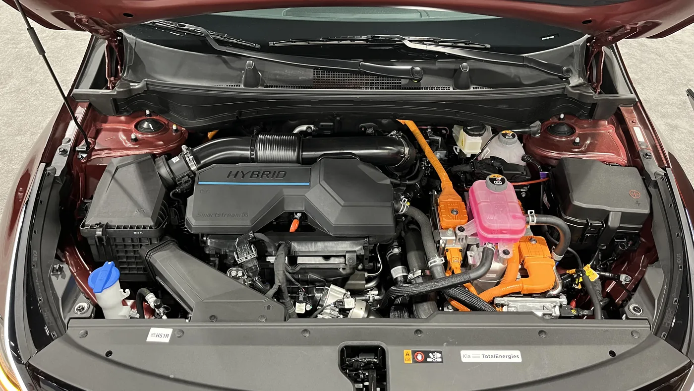 Engine bay of a Kia Sportage Hybrid with the hood open, showing the engine cover, orange high-voltage cables and coolant reservoirs