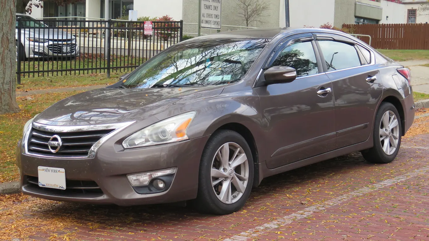 Brown 2015 Nissan Altima sedan parked on a brick street covered with fallen leaves, seen from the front left