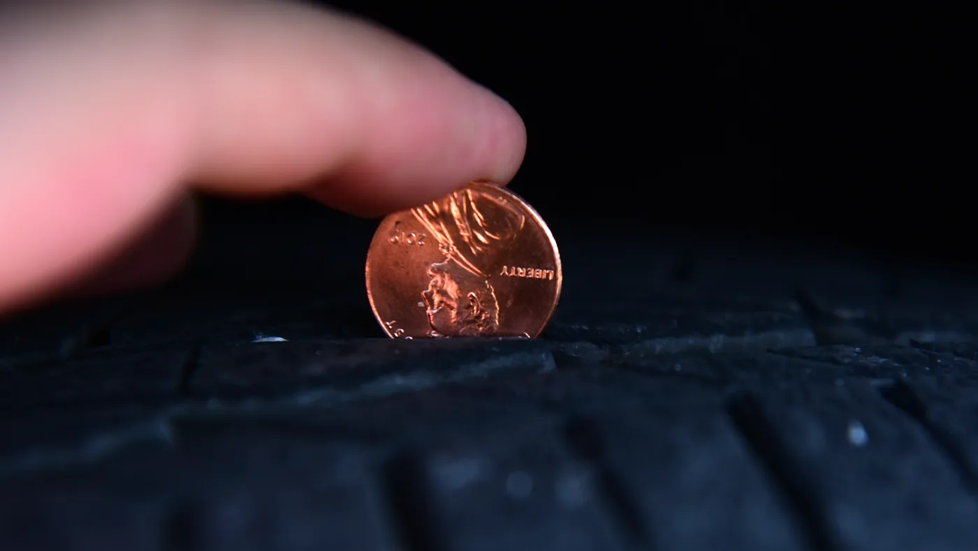 Fingers holding a penny upside down in a tire tread groove to check tread depth against Lincoln's head