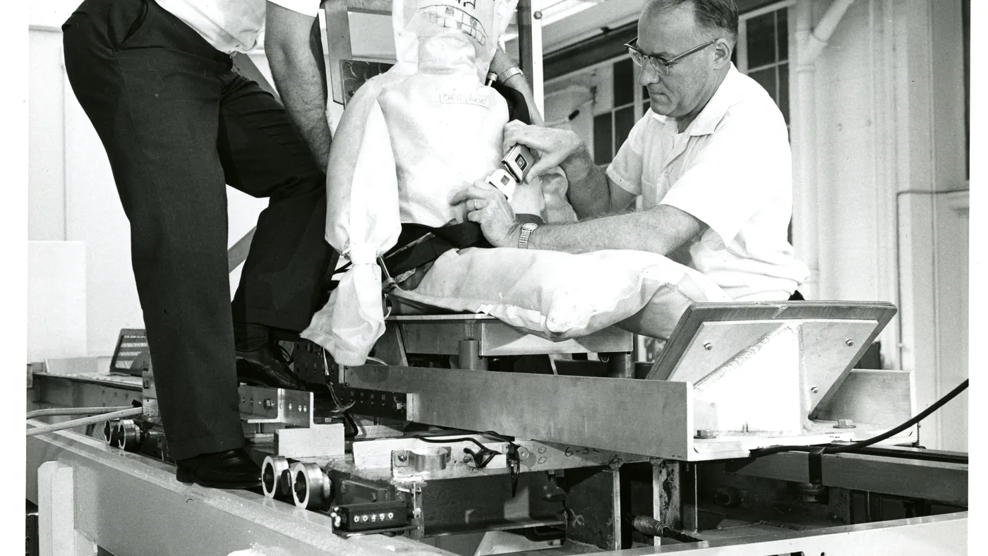 Black-and-white photo of a technician fastening a seat belt across a crash-test dummy seated on a test sled