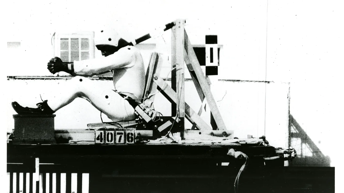 Black-and-white photo of a helmeted crash-test dummy restrained in a seat on a test sled, with a frame number board in front