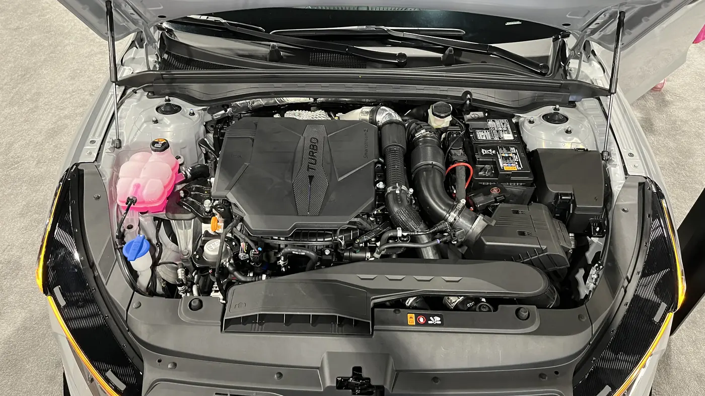 Open hood of a white sedan at an auto show showing the turbocharged engine, coolant reservoir and battery