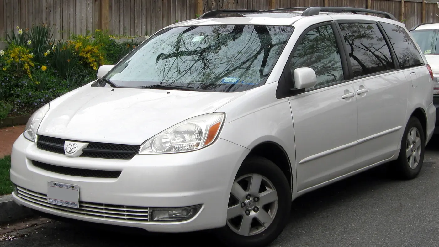 White 2004 Toyota Sienna minivan parked at a curb with spring flowers behind it, seen from the front left